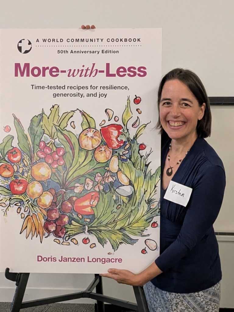 Kirsten poses with a giant cover of the 50th anniversary edition of the More-with-Less, decorated with a cornucopia of abundant produce.